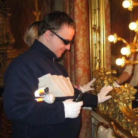 Meżczyzna w ciembych okularach dotyka w rękawiczkach swiecącego kinkietu przy lustrze.