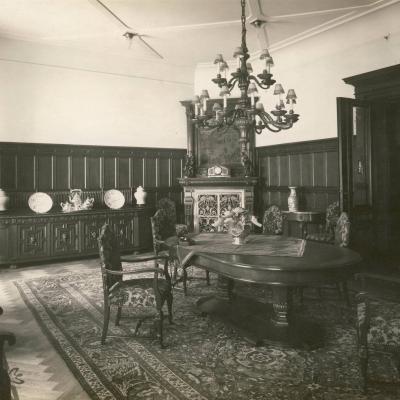 Czarno-białe zdjęcie przedstawia pokój. Na środku pokoju znajduje duży stół. Wokół stolu stoją folele.